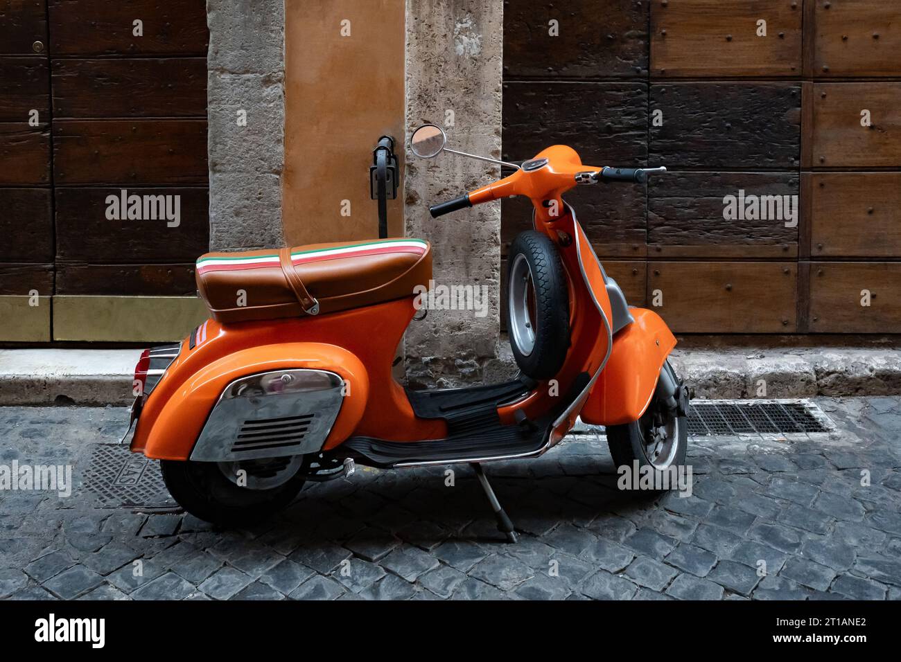 Orange scooter, vespa parked by the wall with wooden doors on the empty