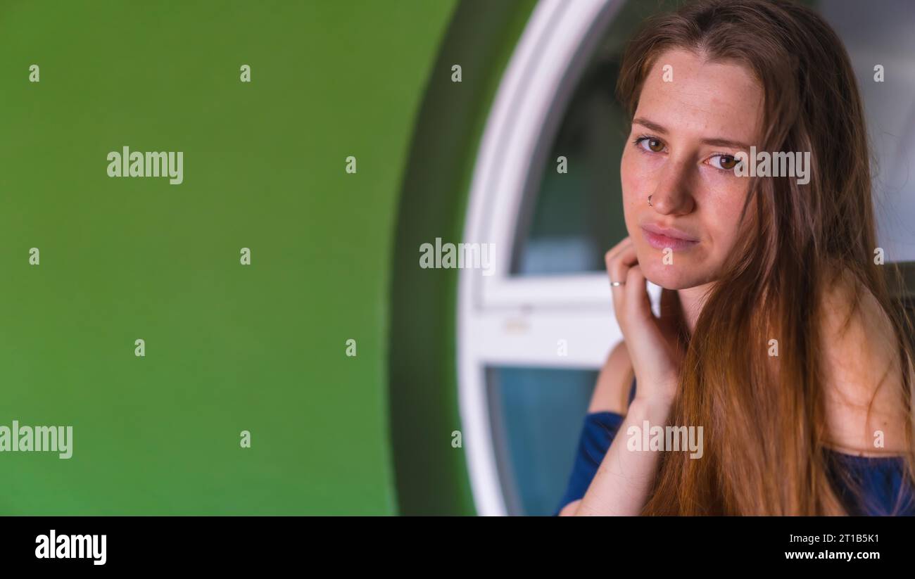 A young pretty red-haired Caucasian woman smiling in a blue dress next to a white sale of a green house, copy space and paste Stock Photo