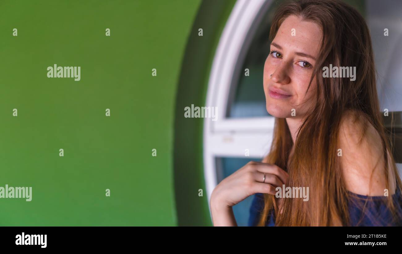 A smiling young pretty redhead Caucasian woman sitting in a blue dress next to a white sale of a green house, copy space and paste Stock Photo