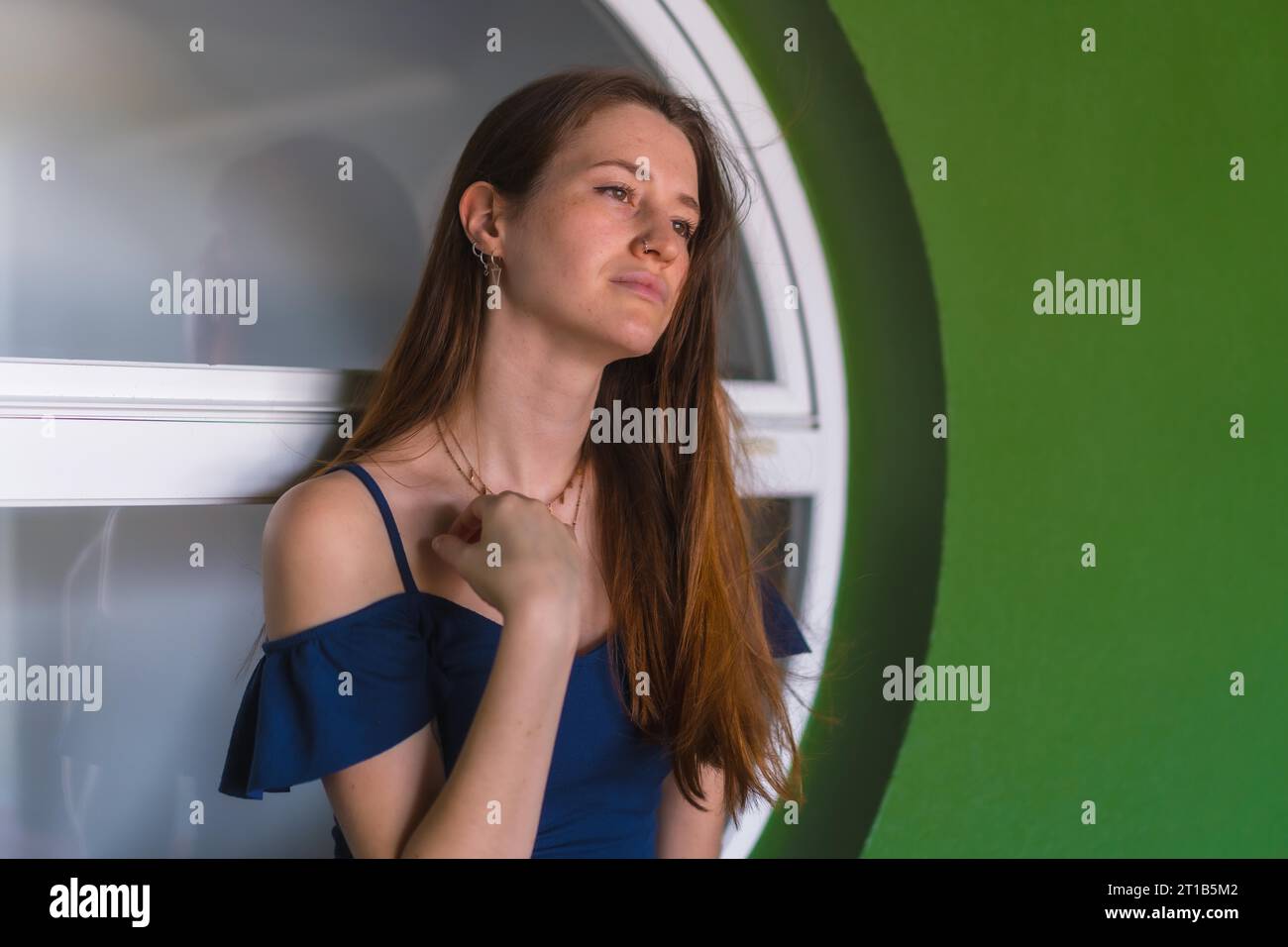 A young pretty redhead Caucasian girl smiling sitting in a blue dress next to a white sale of a green house Stock Photo