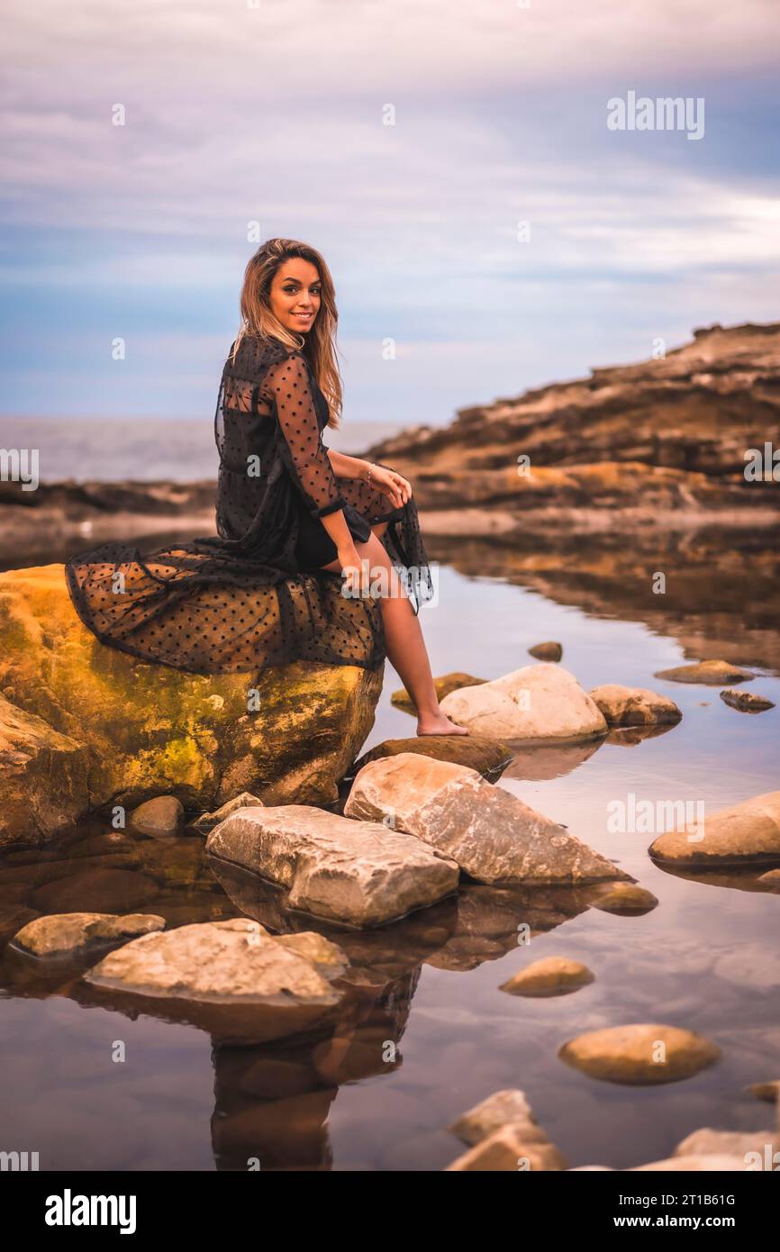 Summer lifestyle with a young brunette Caucasian woman in a long black transparent dress on some rocks near the sea on a summer afternoon. Sitting on Stock Photo