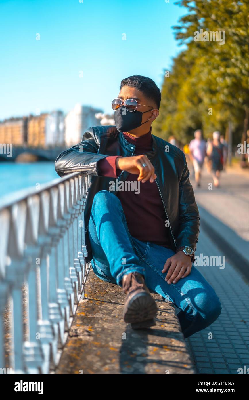 Fashion lifestyle, portrait of a young Latino in the river of the city. Jeans, leather jacket and brown shoes. In a pandemic with a mask Stock Photo