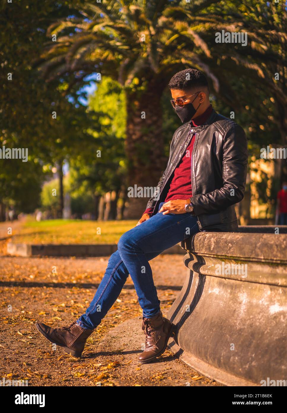 Fashion lifestyle, portrait of a young Latino in the city at a water fountain. Jeans, leather jacket and brown shoes. In a pandemic with a mask Stock Photo