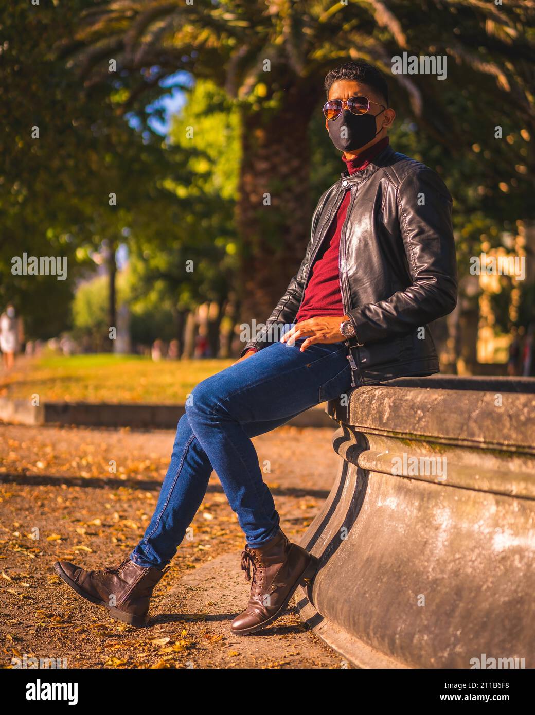 Fashion lifestyle, portrait of a young Latino in the city at a water fountain. Jeans, leather jacket and brown shoes. In a pandemic with a mask Stock Photo