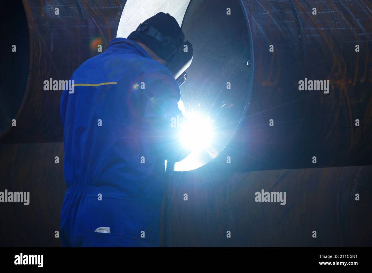 Industrial electrode welder with face shield and blue overall welding a steel pipe in workshop. Back view. Stock Photo