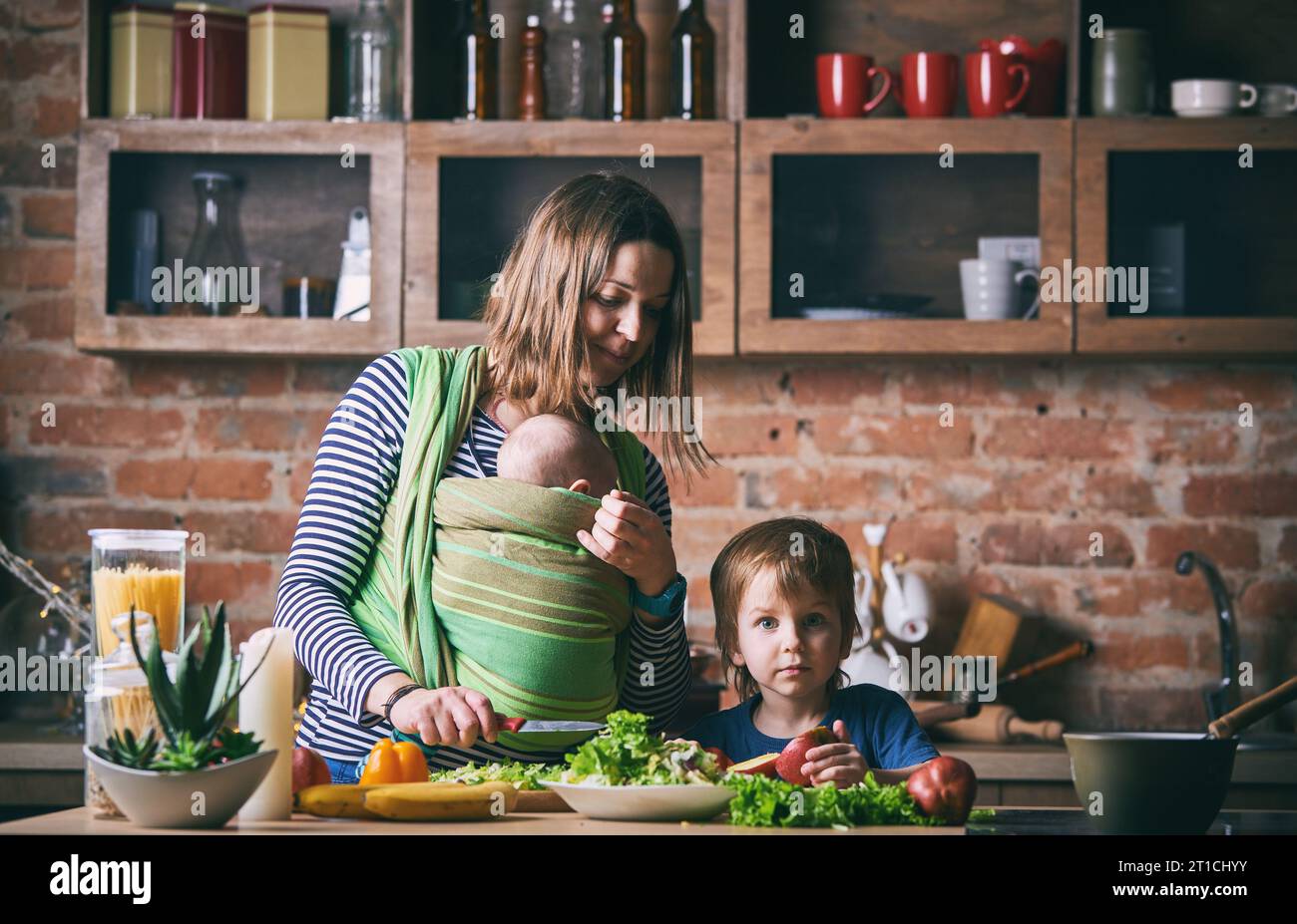 Happy young family, beautiful mother with two children, adorable preschool boy and baby in sling cooking together in a sunny kitchen. Vintage style. Stock Photo