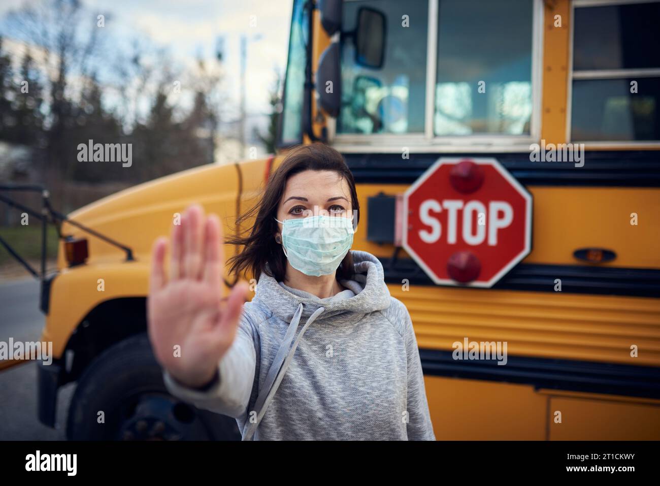A woman in a protective mask stands on the background of a school bus. Raises a hand and stops the epidemic of coronavirus. A large stop sign is visib Stock Photo
