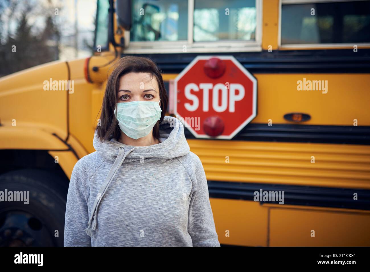 A woman in a protective mask stands on the background of a school bus. A large stop sign is visible on the background. Stock Photo