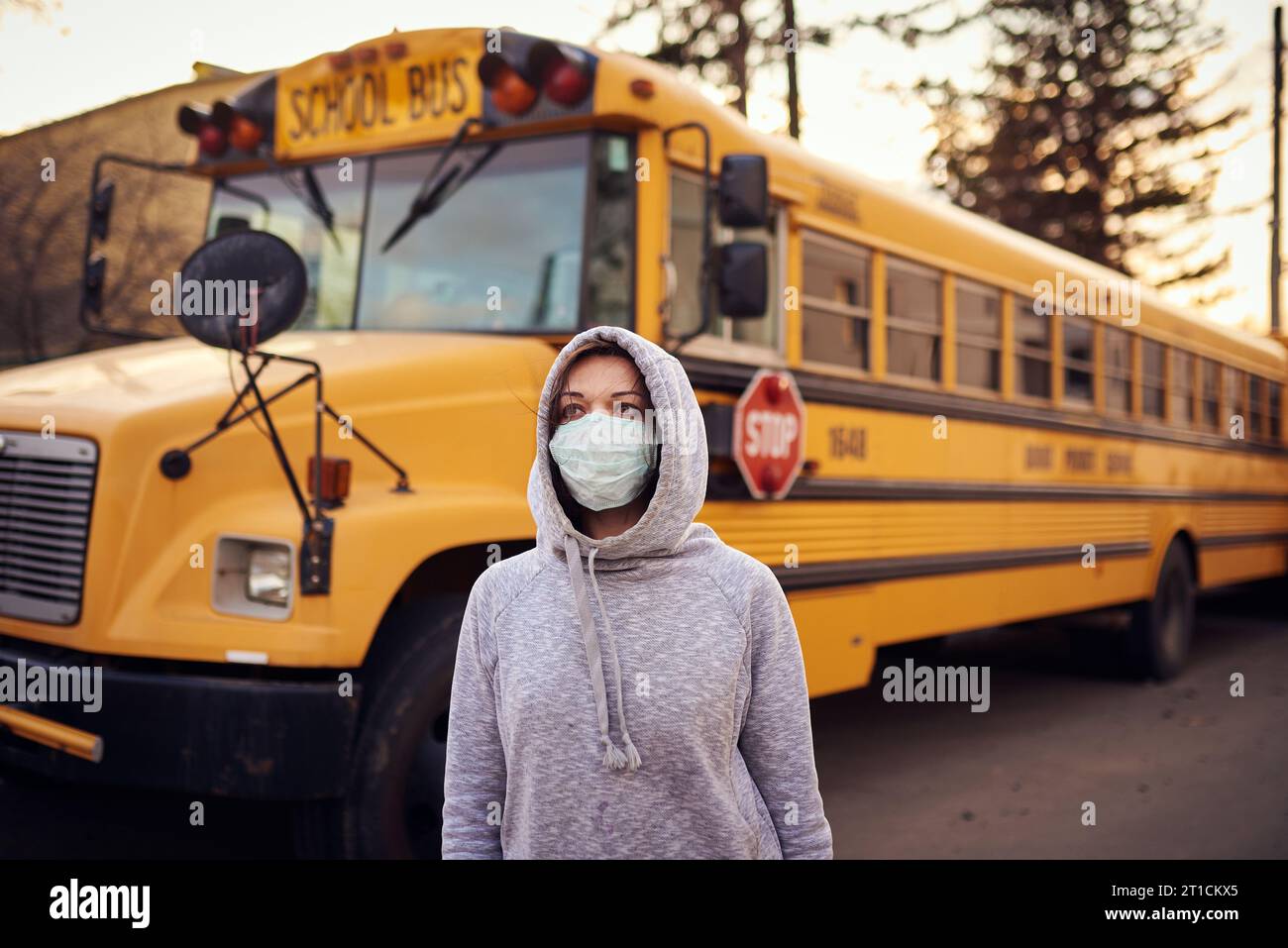 A woman in a protective mask stands on the background of a school bus. A large stop sign is visible on the background. Stock Photo