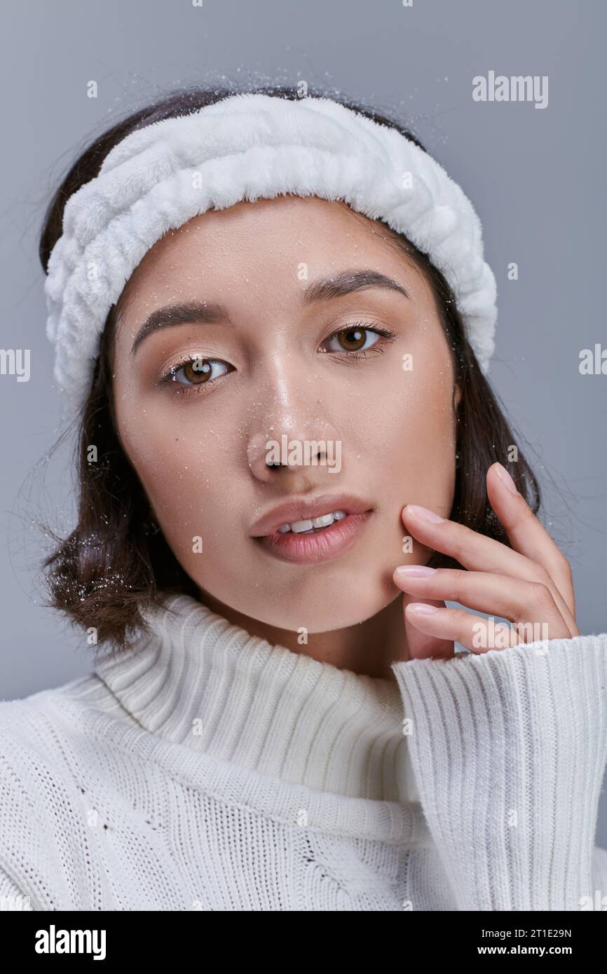 portrait of charming asian woman in warm headband touching face and looking at camera on grey Stock Photo