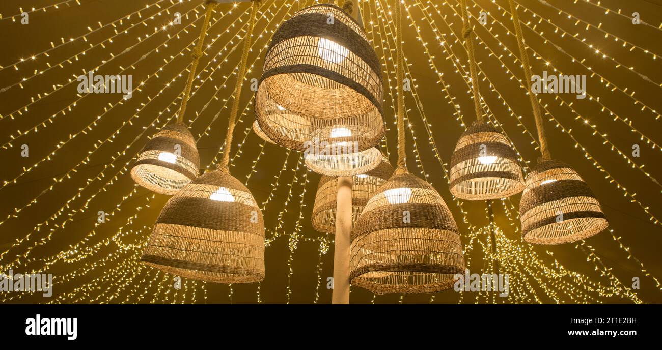 Natural fiber lamps hanging from the ceiling of a tent with a background of LED garlands. Warm atmosphere Stock Photo