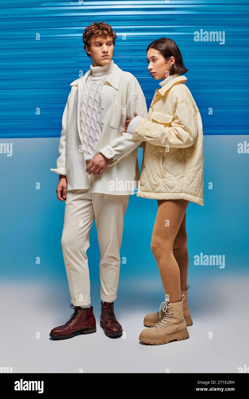 trendy interracial couple in warm outerwear posing on blue backdrop with plastic sheet, winter style Stock Photo