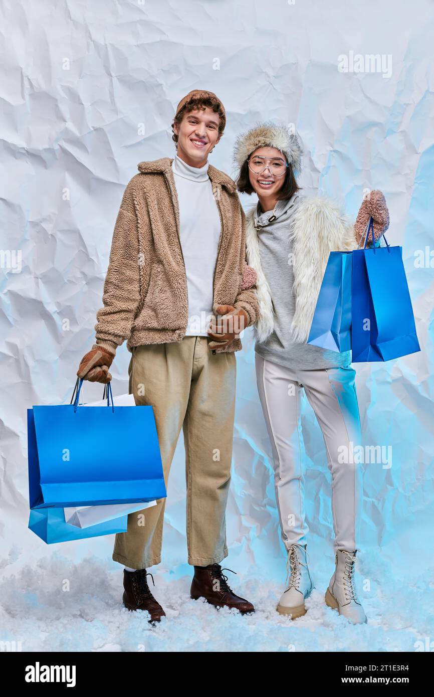 joyful multiethnic models in warm outerwear with blue paper bags on snow in studio, winter shopping Stock Photo