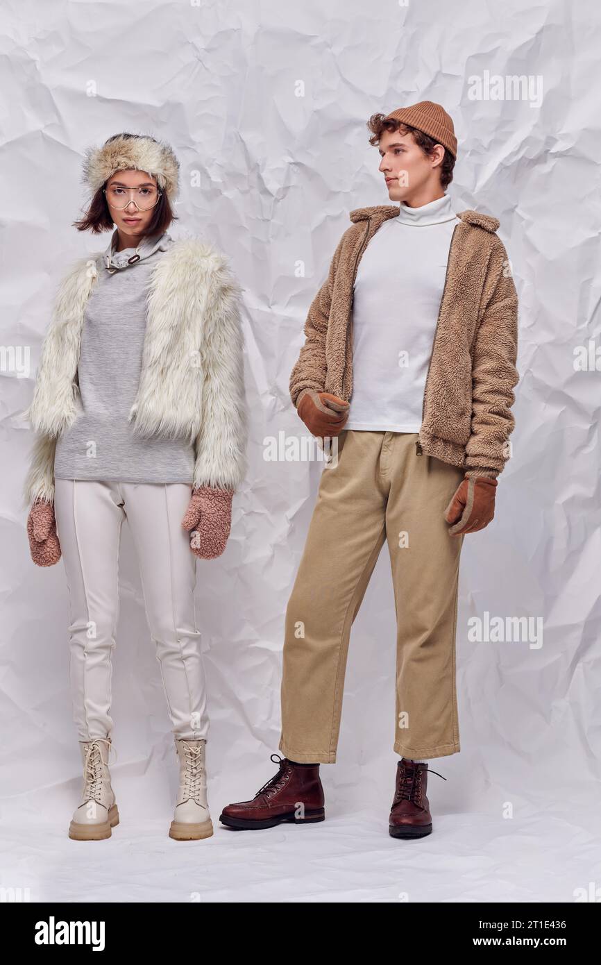 winter fashion campaign, young interracial couple in trendy outerwear on white crumpled backdrop Stock Photo