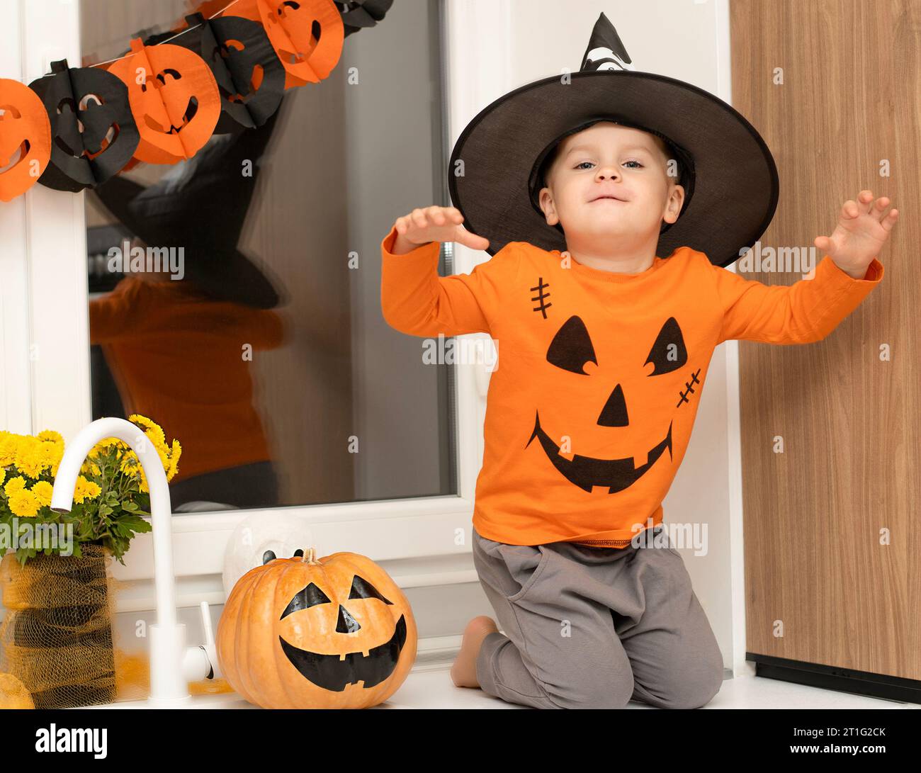 Halloween concept. A small, cheerful, handsome boy in a wizard's hat and an orange sweater sits on a table in the kitchen against the background of a Stock Photo