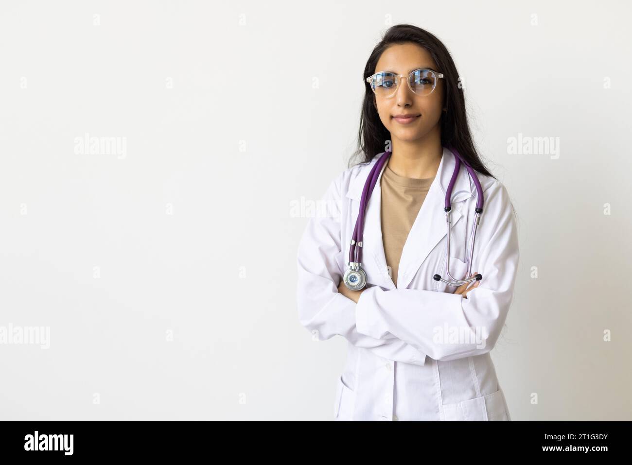 Pretty young smiling clinician in whitecoat crossing arms on chest while looking at you in isolation Stock Photo