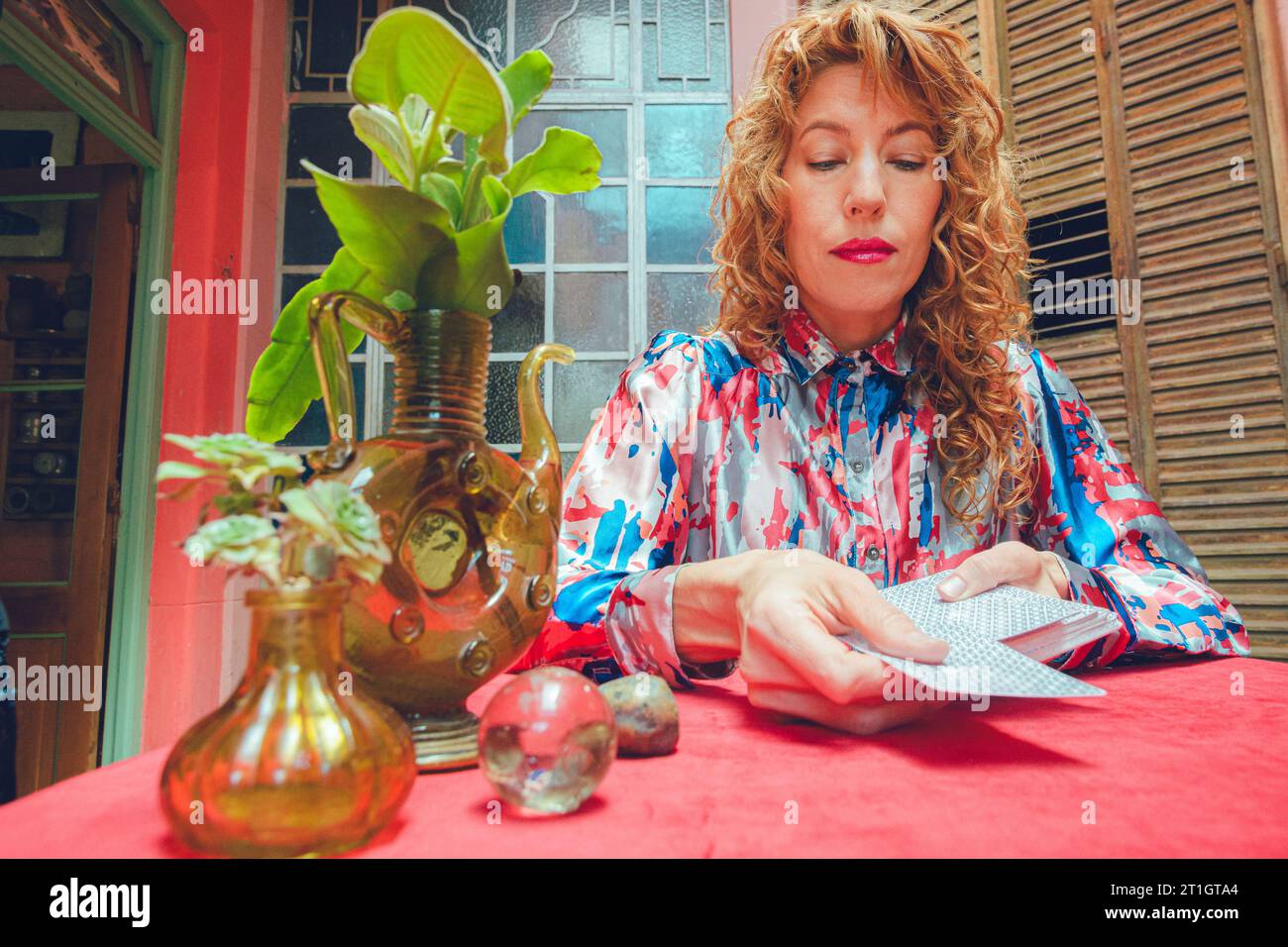 elegant blonde Argentinian adult tarot reader woman with curly hair, sitting at home beginning reading taking card out of deck to place it on table, c Stock Photo