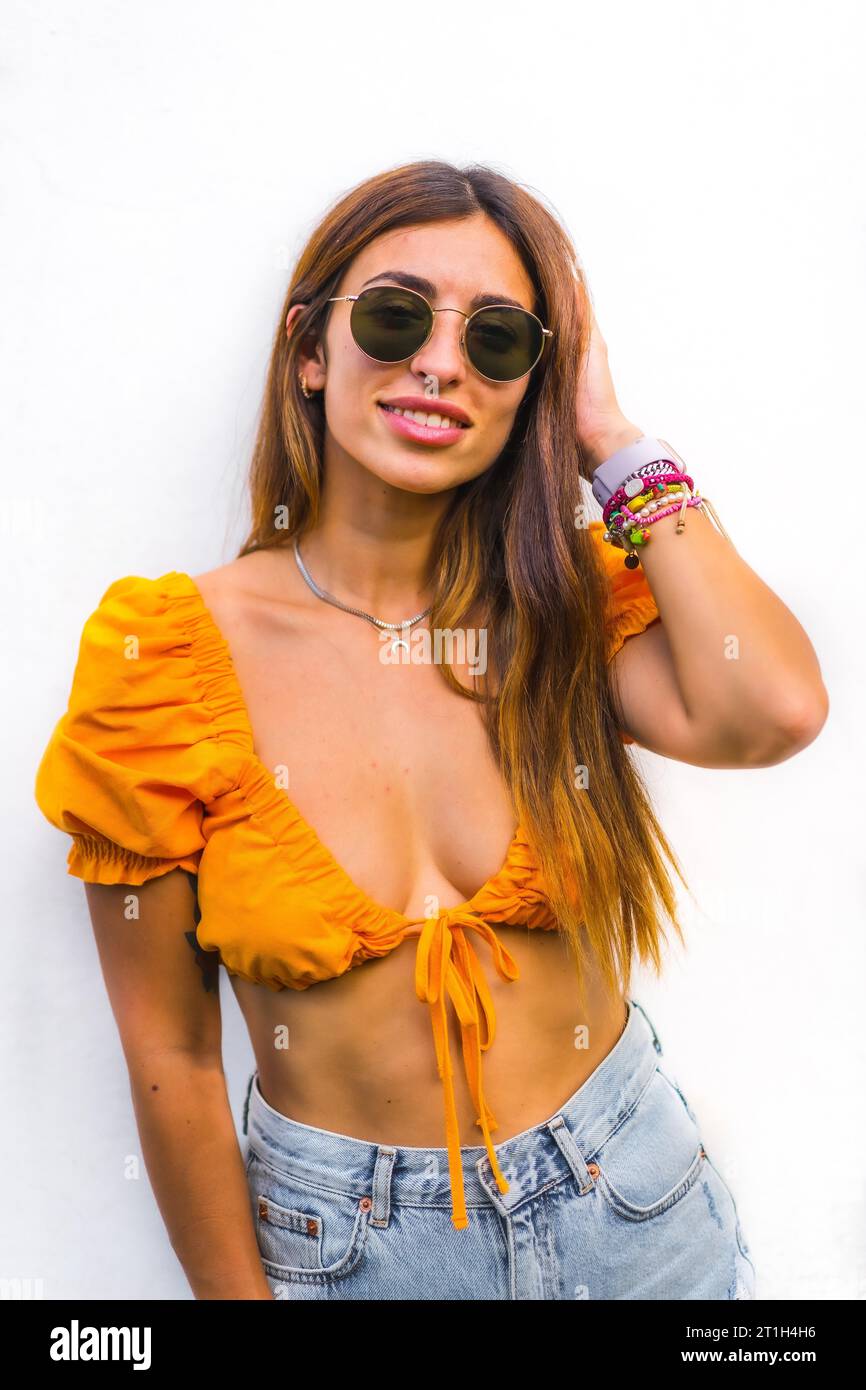 Lifestyle of a young caucasian brunette with orange t-shirt, sunglasses and short pants on a white background, smiling looking at camera Stock Photo