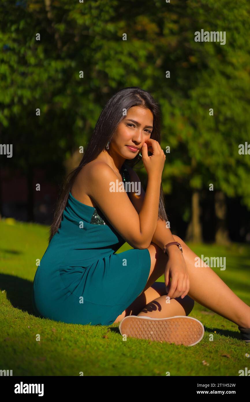 A pretty young brunette Latina with long straight hair, wearing a tight green dress. Portrait of a sitting on the grass in a park in summer Stock Photo