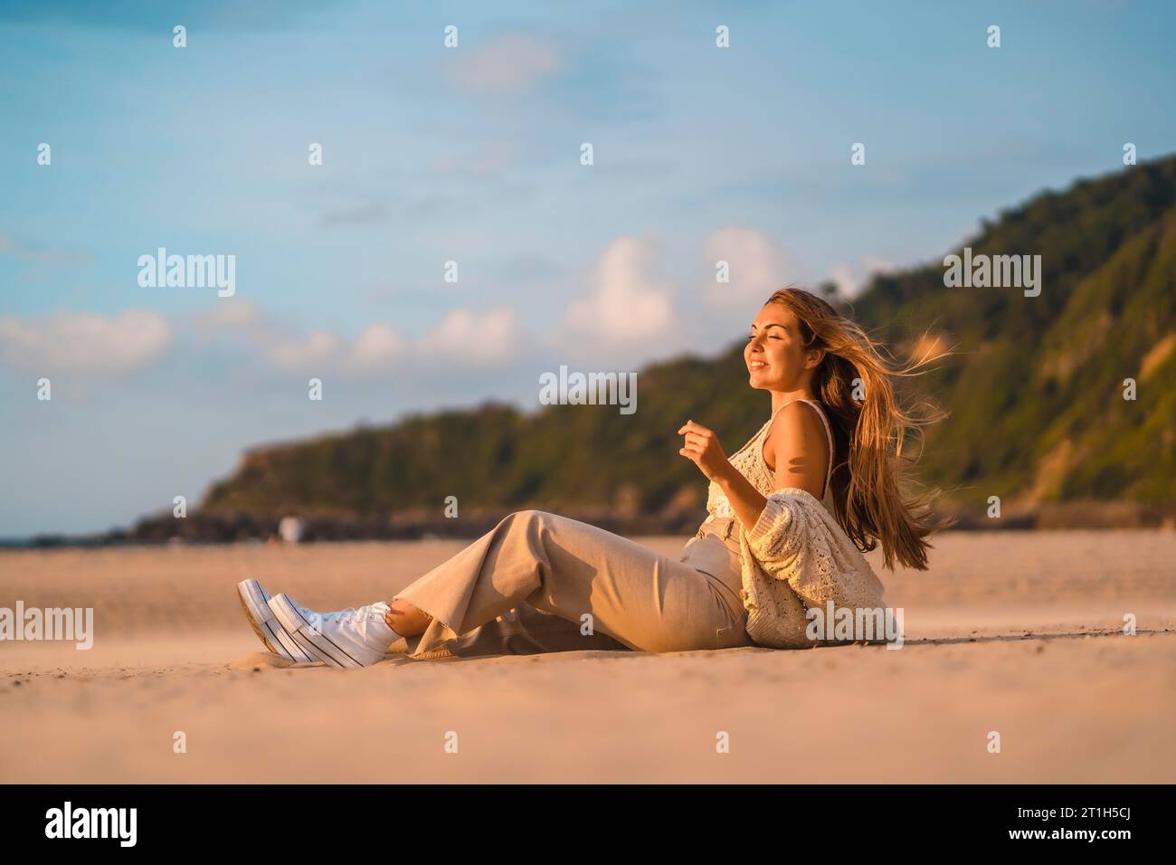 Summer lifestyle, a young blonde with straight hair, in a small wool sweater and corduroy pants sitting on the beach with mountains and the sea in Stock Photo