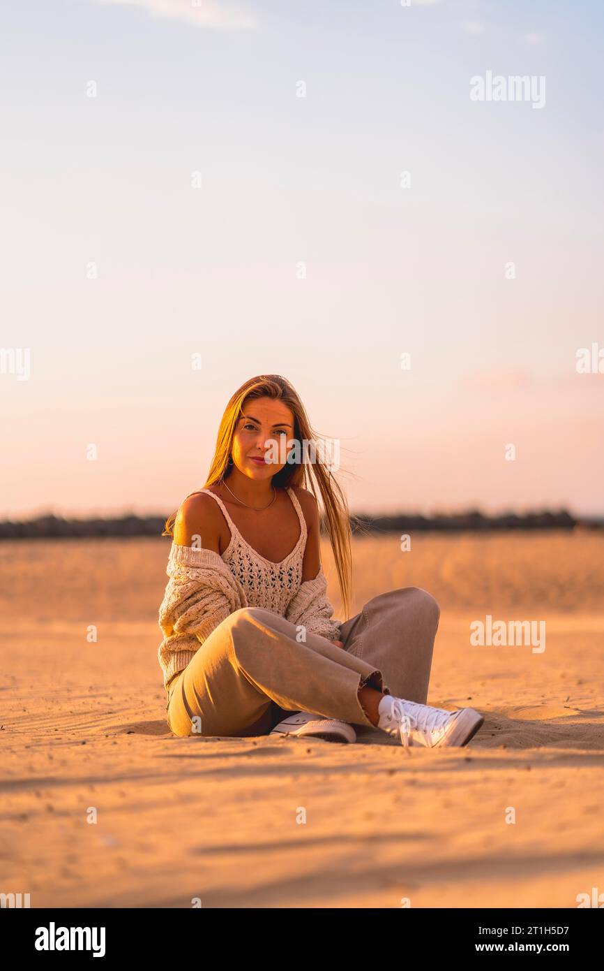Summer lifestyle, a young blonde with straight hair, in a small wool sweater and corduroy pants sitting on the beach. Sitting on the sand smiling a Stock Photo