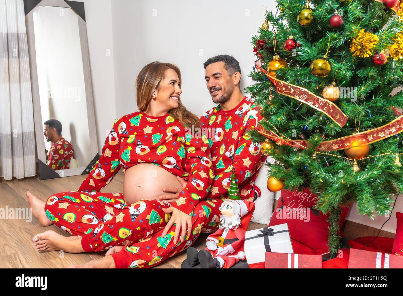 Young couple smiling sitting next to the Christmas tree with gifts. Family with pregnant woman Stock Photo