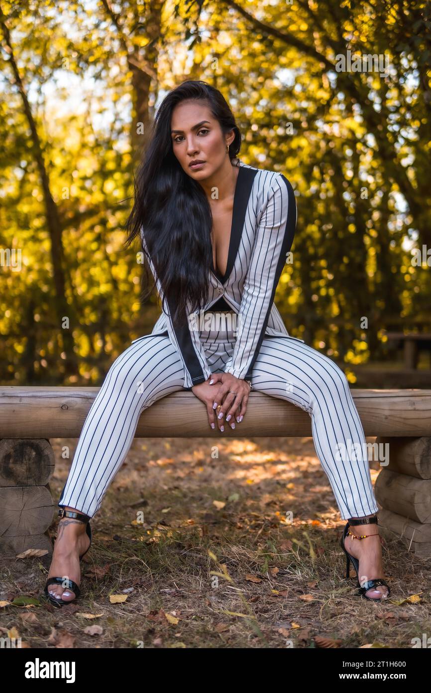 Summer lifestyle, young Latin brunette girl in a very elegant white suit with black stripes. Fashion posed in a beautiful park sitting, fashion Stock Photo