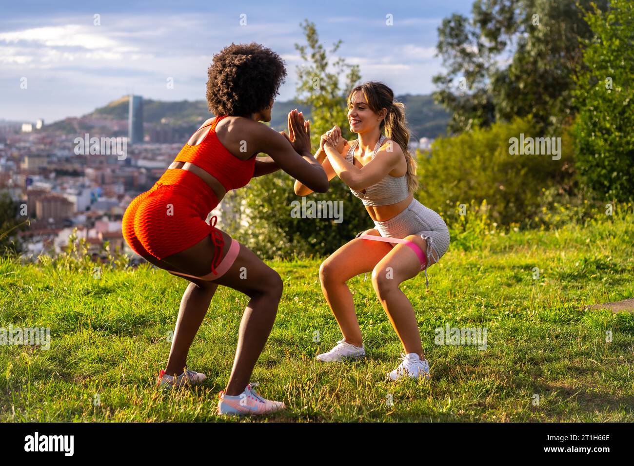 Caucasian blonde girl and dark-skinned girl with afro hair doing squat exercises in a park with the city in the background. Healthy life, fitness Stock Photo