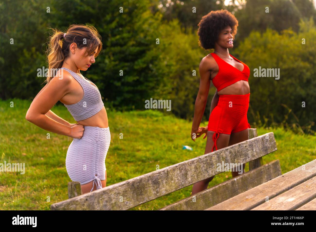 Caucasian blonde girl and dark-skinned girl with afro hair doing stretching before starting sports in the park. Healthy life, fitness, fitness girls Stock Photo