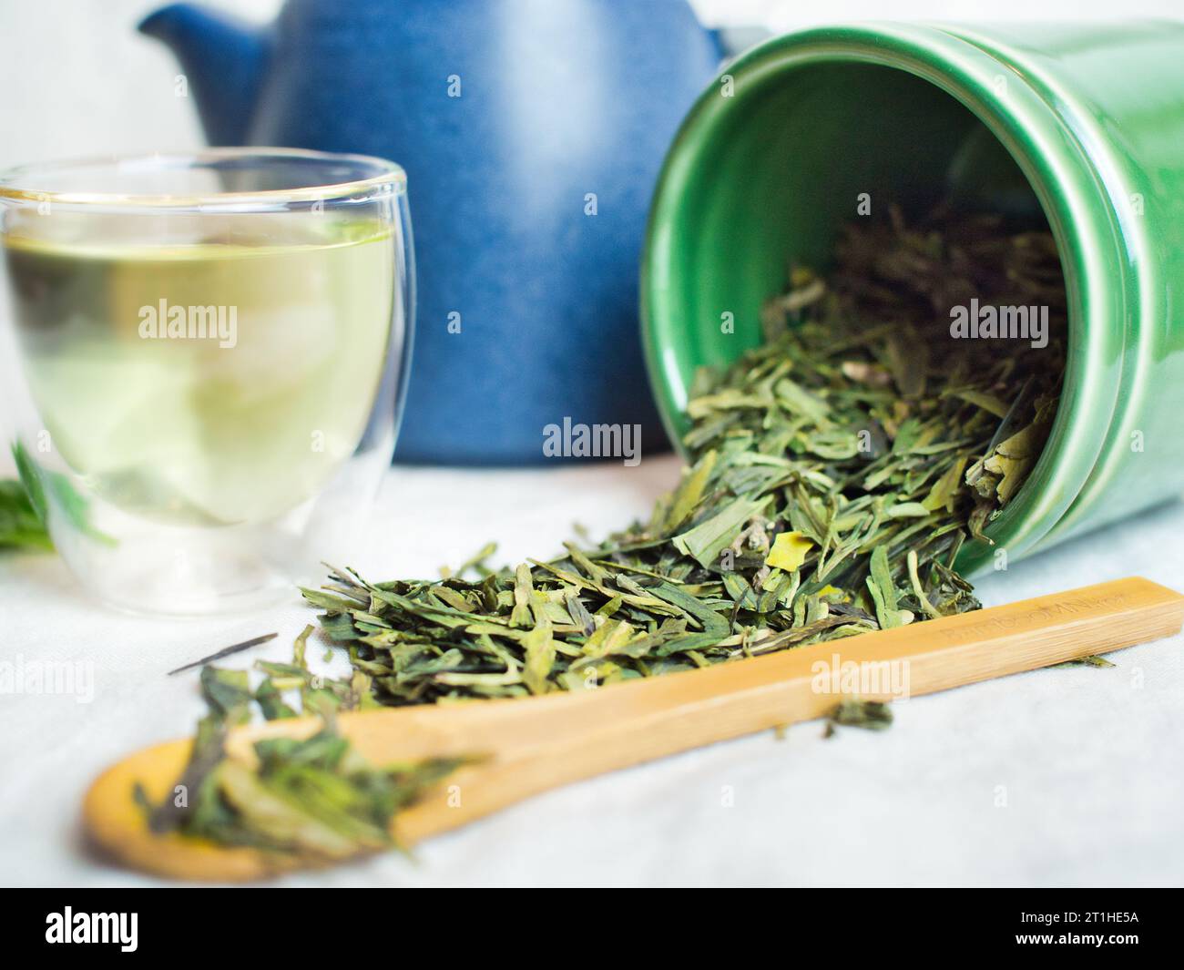 Still life of green tea long jing dragonwell from China. Tea leaves shown on linen table cloth with green tea in cup, teapot, and bamboo spoon. Stock Photo