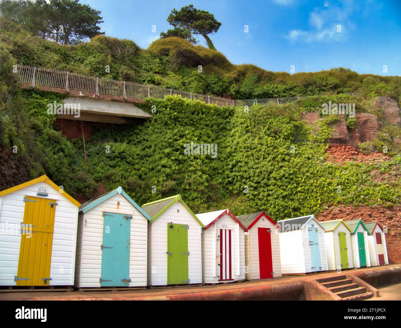 A row of beach huts with their colourful doors on the promenade of Goodrington North Sands below the cliff walk of Roundham Head, Paignton. Stock Photo