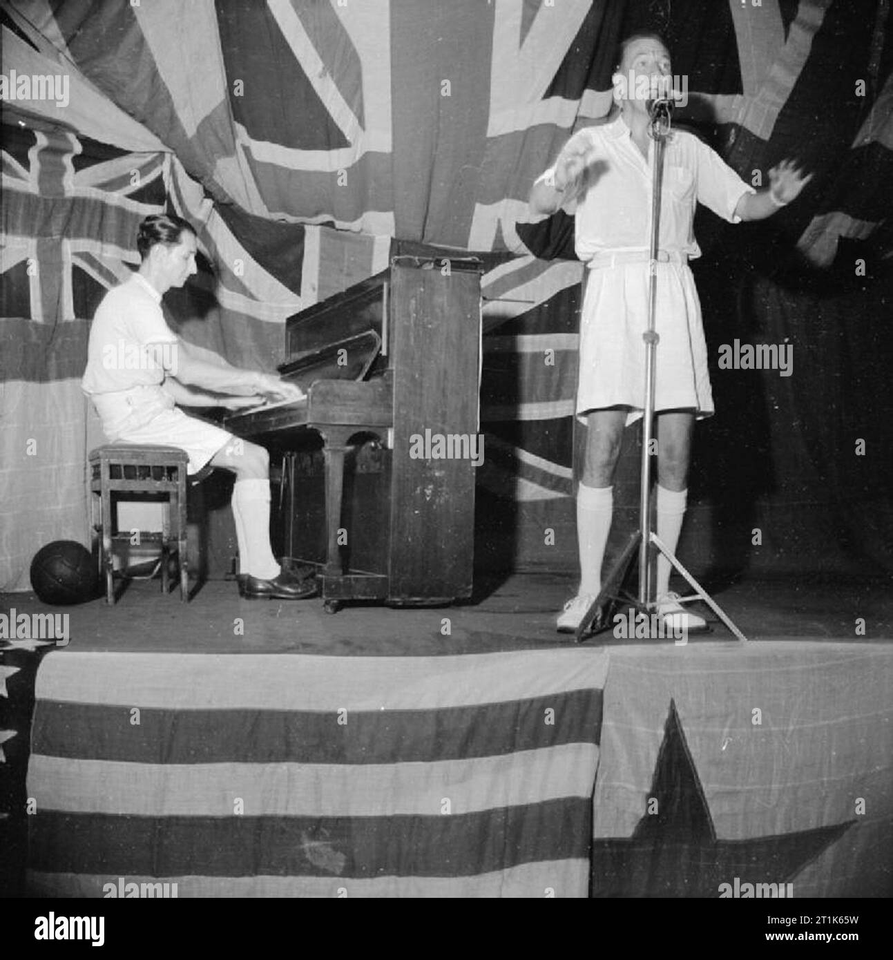 Noel Coward Entertains the Men of the Eastern Fleet, HMS Victorious, Trincomalee, Ceylon, 1 August 1944 Noel Coward standing at the microphone on a flag-bedecked stage on the aircraft lift aboard HMS VICTORIOUS with Norman Hackforth at the piano. Stock Photo