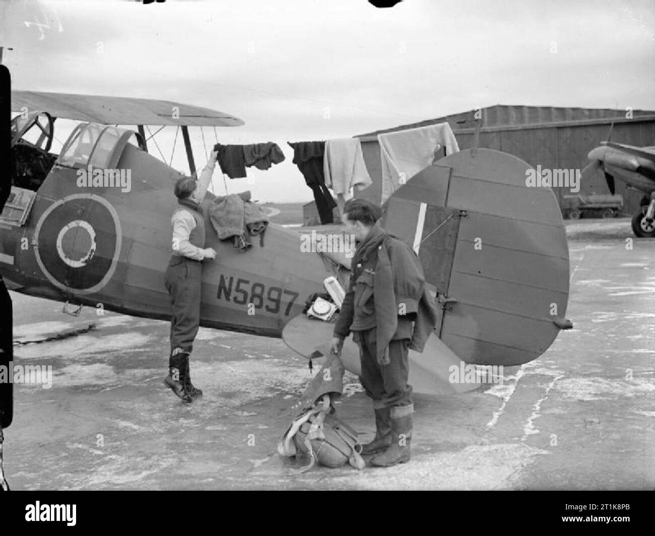 Royal Air Force Coastal Command, 1939-1945. A meteorological pilot, Flying Officer J B Gordon of No. 521 Squadron RAF, prepares to don the many layers of clothes he wears for high-altitude weather flying, which are hanging from the radio aerial of his Gloster Gladiator Mark II, N5897 'E', at Bircham Newton, Norfolk. A ground crewman stands by with Gordon's SD jacket and parachute. A psychrometer and an aneroid barometer, which will be attached to the aircraft for the THUM (Temperature and Humidity) flight, have been placed on the elevator. Stock Photo