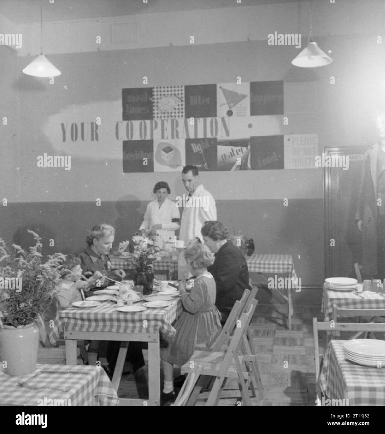Civil Defence and Welfare Services in Wartime Britain- the work of the Rest Centre, 1942 A bombed-out family enjoy a meal at a rest centre. Checked table cloths and flowers provide a welcoming and cheery atmosphere to those who have lost their homes through air raids. One of the two welfare workers visible in the photographs brings a cup of tea over to the table. Stock Photo