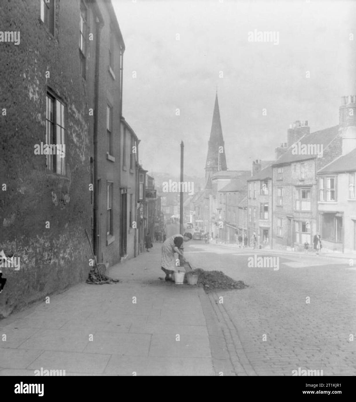 A Middle East Soldier Revisits Britain- Life in Wartime Durham, England, UK, 1943 A woman uses several buckets to collect coal from a large pile outside the houses in Claypath, Durham. This photograph was taken from the top of Claypath, looking down the almost completely deserted street, which slopes away to the west. The spire of Claypath Congregational church can be seen in the background. Stock Photo
