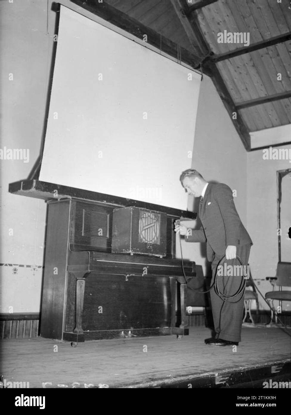 Film Show at Highest Village in the Highlands- Ministry of Information Film Screening, Tomintoul, Banffshire, Scotland, UK, 1943 Ministry of Information Film Operator John Macdougall sets up a screen and loudspeaker in the Memorial Hall in Tomintoul in preparation for the screening of a Ministry of Information film. The loud speaker is resting on the closed lid of an upright piano, with the screen resting on the top of the piano. Mr Macdougall is connecting the 'soundbox'. The original caption states that 'power is provided by the generator in [his] van outside'. Stock Photo