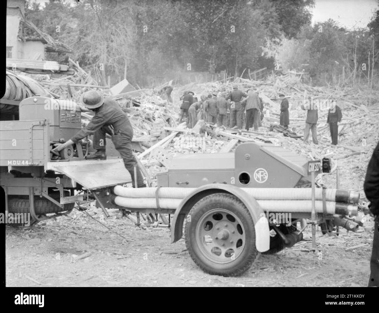 Flying Bomb- V1 Bomb Damage in London, England, UK, 1944 A fireman of the National Fire Service prepares to lift equipment from a truck following a V1 attack in the Highland Road and Lunham Road area of Upper Norwood. An NFS trailer pump can be seen in the foreground, standing ready should it be required. In the background, a group of Civil Defence rescue workers search for survivors under a huge pile of rubble and timbers. Stock Photo