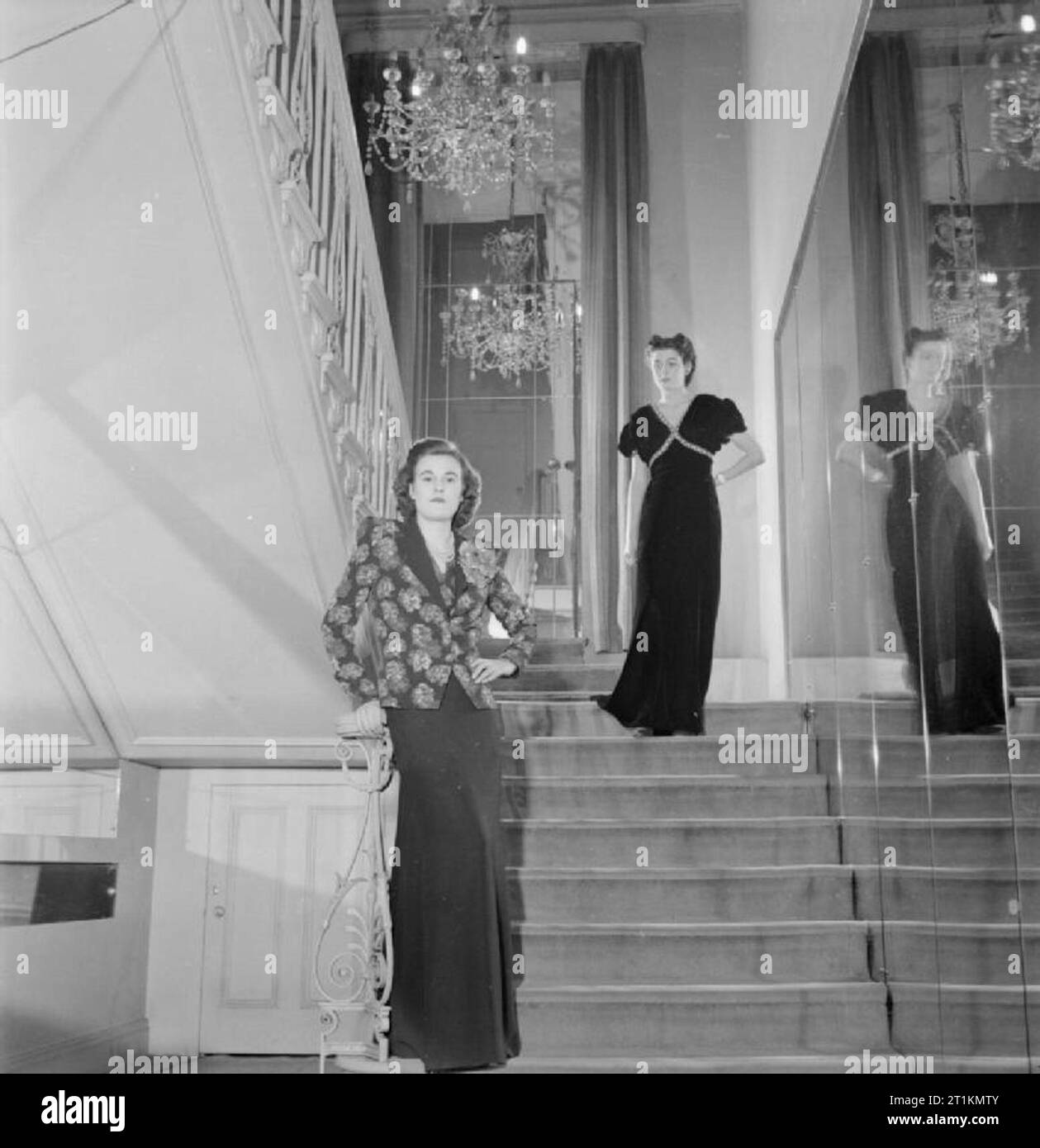 London Fashion Designers- the work of Members of the Incorporated Society of London Fashion Designers in Wartime, London, England, UK, 1944 Two models pose for the camera on a staircase at the fashion house of designer Norman Hartnell. At the top of the stairs, the model wears a black velvet dinner gown studded in blue and gold. In the foreground, the model wears a dinner suit with the jacket made from a red rose printed fabric, designed by Hartnell and a skirt of black crepe. A large chandelier is clearly visible in the background. Stock Photo