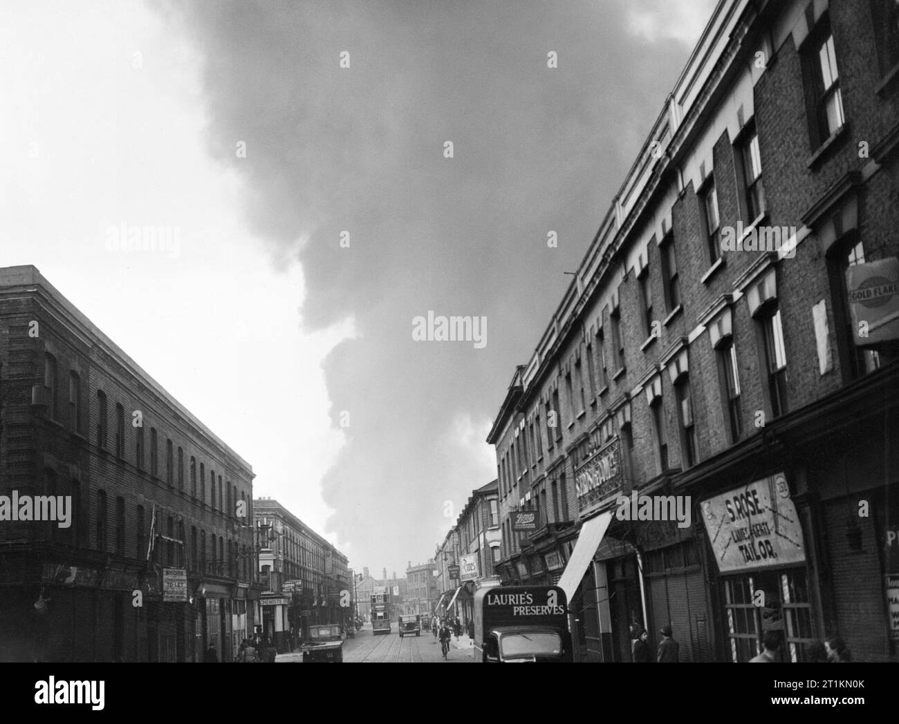 A cloud of smoke rises in the distance after a V1 Flying Bomb lands in the Norwood area of London during 1944. A cloud of smoke rises in the distance after a V1 attack in the Norwood area of London. Life appear to be continuing as normal, however, as this busy high street scene illustrates. At the far end of the street (possibly Norwood High Street or Lower Addiscombe Road), a number 34 tram can just be seen. On the right hand side of the street, shops include 'Old Times Furnishing Company', Boots the Chemist and S Rose's tailor's shop (at number 252). A van delivering 'Laurie's Preserves' can Stock Photo