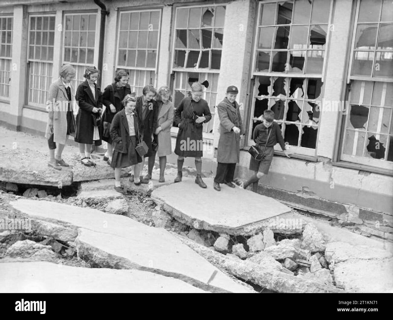 London Schools in Wartime- School Life in London, England, 1941 School children inspect bomb damage in the playground of Moorside Road School, Grove Park, London. One boy is pointing to a large hole in the concrete of the playground. Several of the windows have been smashed. All the boys are carrying their gas masks. Stock Photo