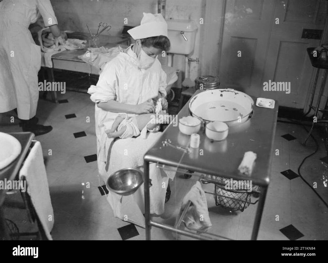 Mansion Becomes Maternity Home- Life at Brocket Hall, Welwyn, Hertfordshire, 1942 A student nurse prepares a baby for a bath at Brocket Hall. The nurse is wearing a surgical mask to prevent the spread of germs to the baby. Stock Photo