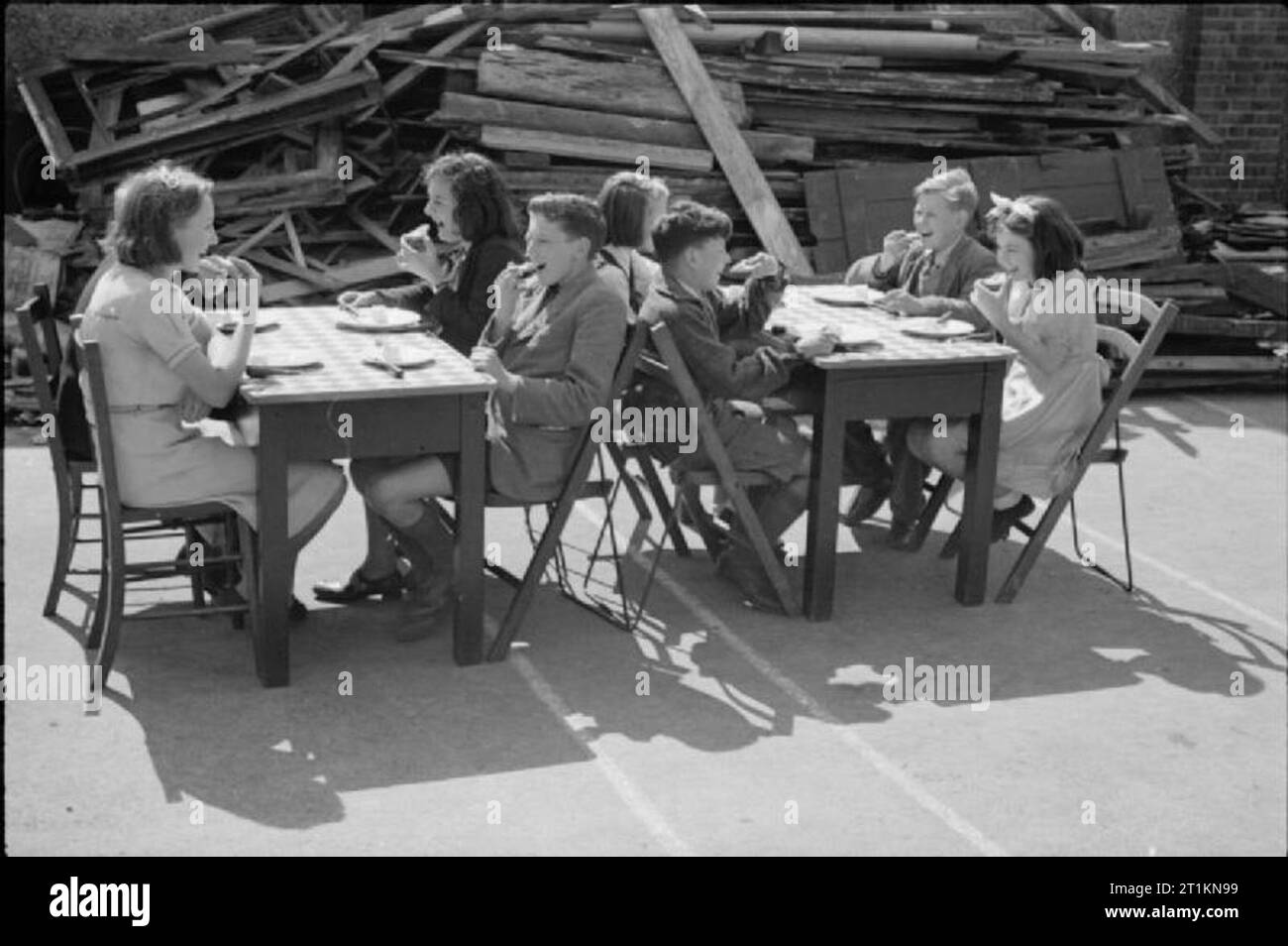 Aid From America- Lend-lease Food, London, England, 1941 Two groups of smiling children sit at tables and eat cheese, imported from America as part of the Lend-Lease scheme, in the playground of a severely bomb-damaged school in London, now being used as a feeding centre. Piles of salvaged wood, including window and door frames, can be seen behind them. This photograph was probably taken in late August or early September 1941. Stock Photo