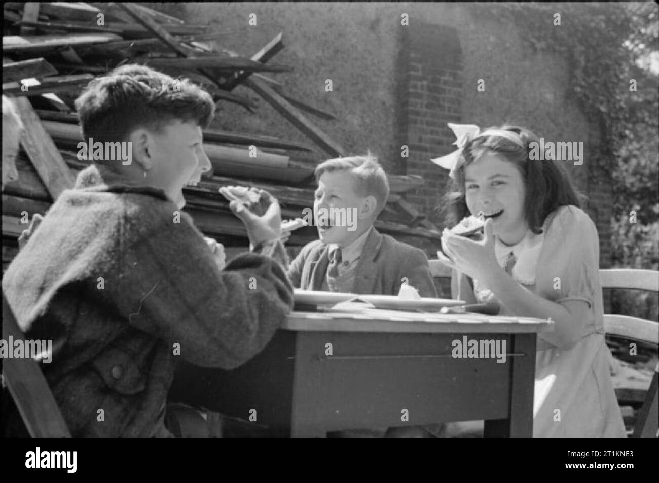 Aid From America- Lend-lease Food, London, England, 1941 A group of smiling children sit at a table and eat cheese imported from America as part of the Lend-Lease scheme in the playground of a severely bomb-damaged school. The school is now being used as a feeding centre. Piles of salvaged wood can be seen just behind the children. This photograph was probably taken in late August or early September 1941. Stock Photo