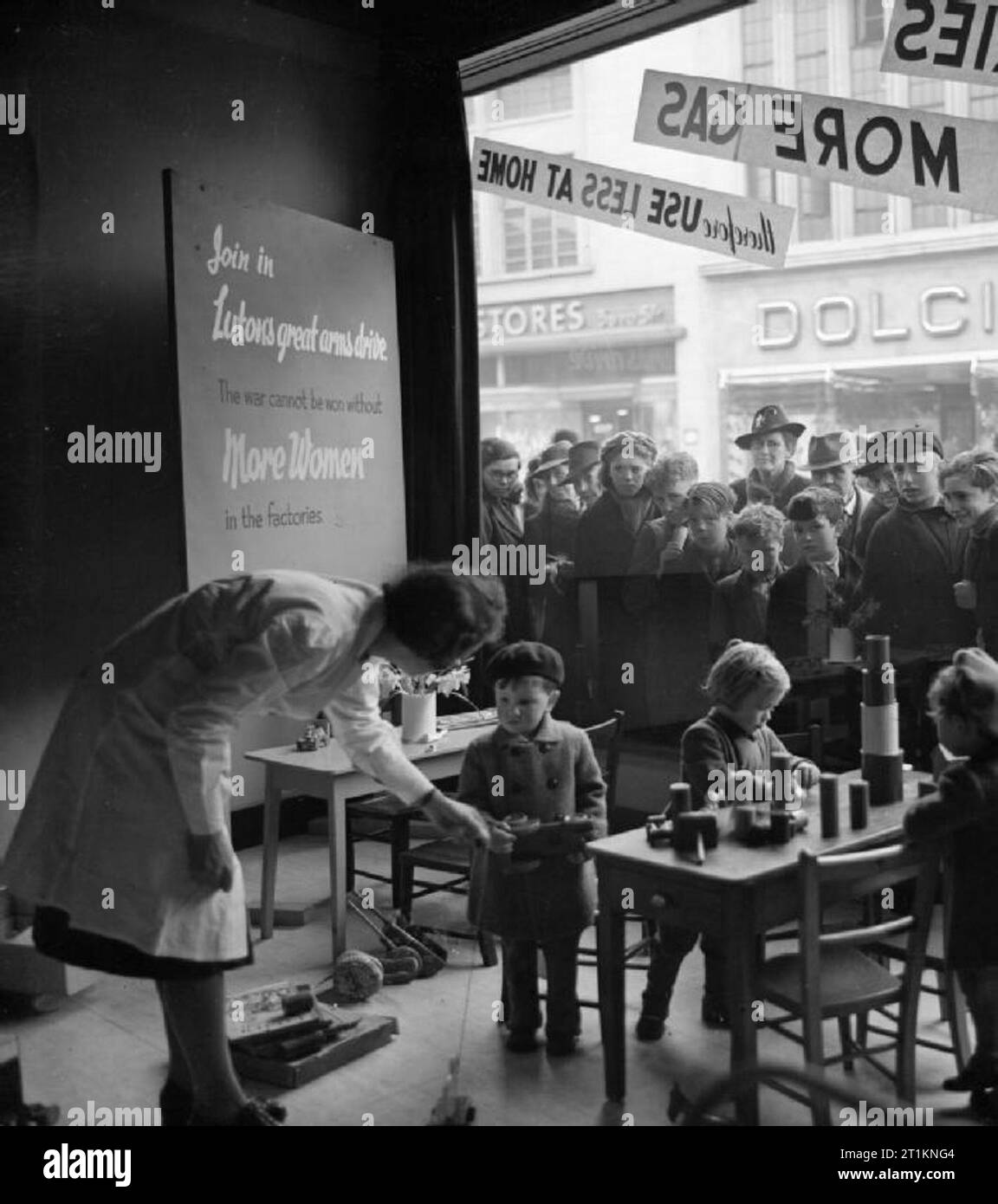 Ministry of Information Exhibitions during the Second World War, Luton, Bedfordshire, England, UK, 1942 A corner of the Women's War Work exhibition in Luton. In the shop window, children play, as a group of women and older children crowd outside to look in at them through the window. A sign reads: 'Join in Luton's great arms drive. The war cannot be won without more women in the factories'. Stock Photo
