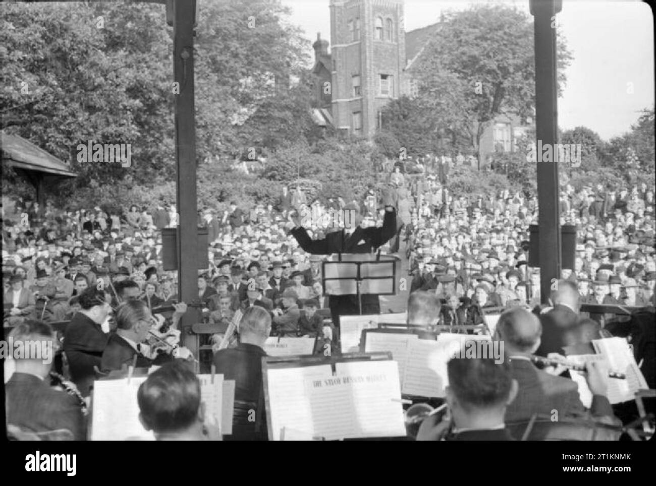 Music Goes To War- Entertainment in Wartime London, England, UK, 1943 Jack Gold conducts his Variety Orchestra in 'The Savoy Russian Medley' on the bandstand in the Horniman Gardens, Forest Hill. The musicians can be seen in the foreground, including those playing the flute, trumpet, violin and clarinet. In the background, the crowd can be seen, enjoying the music in the evening sunshine. Stock Photo