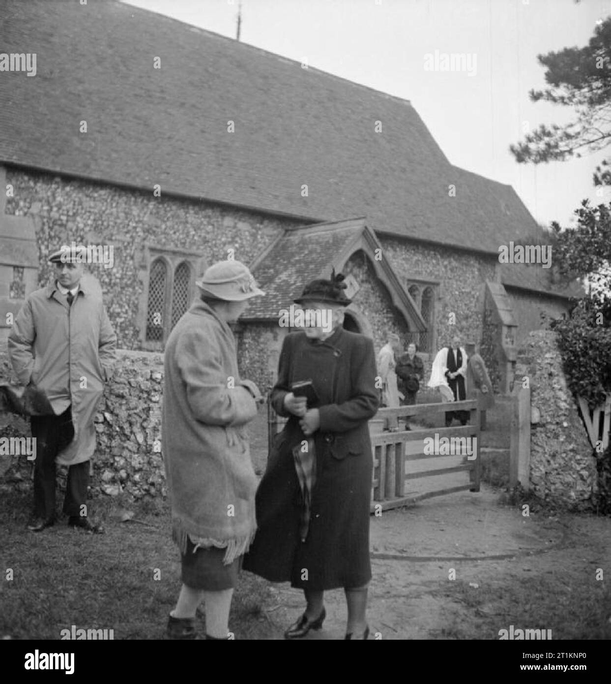 Mrs Bugler Goes To War- Everyday Life in East Dean, Sussex, England, 1943 Mrs Bugler chats to a friend outside the Church of St Simon and St Jude, East Dean, after the Sunday service. In the background, the vicar, Reverend W E N Williams, can be seeing talking to other parishioners. Mrs Bugler's late husband is buried in this churchyard. Stock Photo