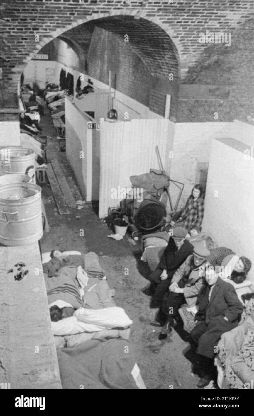 Air Raid Shelter Under the Railway Arches, South East London, England, 1940 A wide view of one of the passages connecting the main sleeping areas at an air raid shelter under the railway arches, in November 1940. Sections of the space have been partitioned off by blast walls. Many shelterers are having to sit or sleep on wooden platforms which have been erected here in the passageway due to overcrowding in the main areas of the shelter. In the background, the gentlemen's lavatories can be seen. Stock Photo