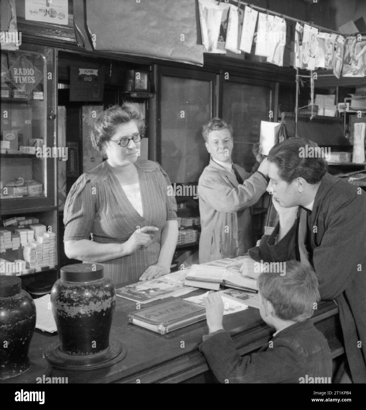 Parish Priest- the work of the Vicar of St Mark's Church, Victoria Docks, Silvertown, London, England, UK, 1944 Reverend Joseph Stephens runs through the music for Sunday service with newsagent and church organist Mrs Fry in her shop in Silvertown. A small boy stands next to Rev Stephens as he waits to buy stamps from Mr Fry, who is working behind the counter with his wife. Stock Photo