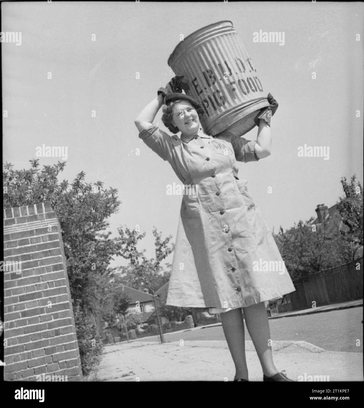Pig Food- Women's Voluntary Service Collects Salvaged Kitchen Waste, East Barnet, Hertfordshire, England, 1943 A full-length portrait of Kathleen Kent, carrying a pig bin on her shoulder, on a sunny street during her daily rounds. Kathleen is the head of the pig food collection unit at East Barnet. Stock Photo