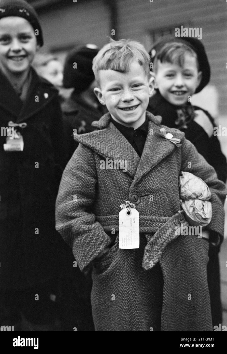 A Dutch refugee arrives at Tilbury in Essex during 1945. A small Dutch boy smiles for the camera upon arrival at Tilbury in Essex. He is carrying a small paper parcel under his arm, which contains all his luggage. He, and the other children, (some of whom can be seen behind him) all have labels pinned to their coats which bear their names, home address and destination. Stock Photo